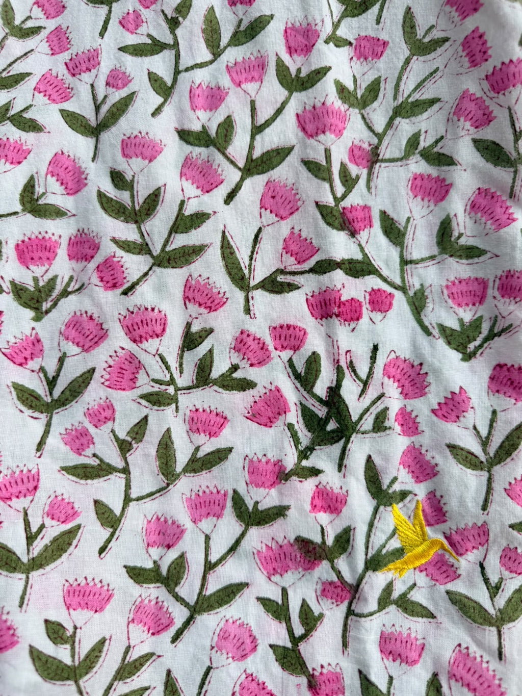 A close-up of a white fabric with a repeating hand-block print pattern of pink flowers and green leaves, featuring a small yellow flower accent.